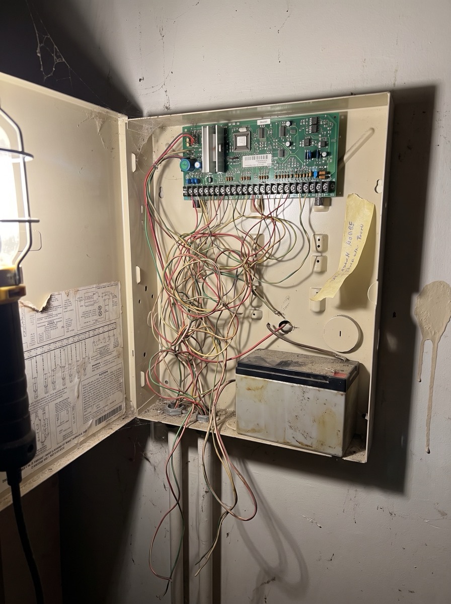 An old alarm panel found during a survey: tangled unlabeled wiring, a corroded battery and dust