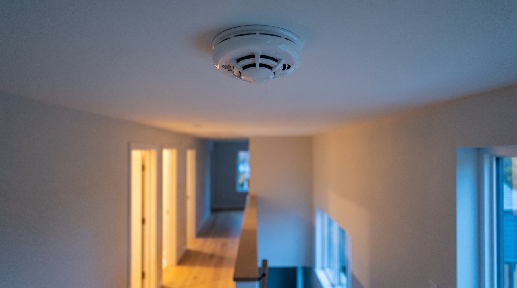 A ceiling-mounted smoke and heat detector above a quiet, empty upstairs landing in the evening