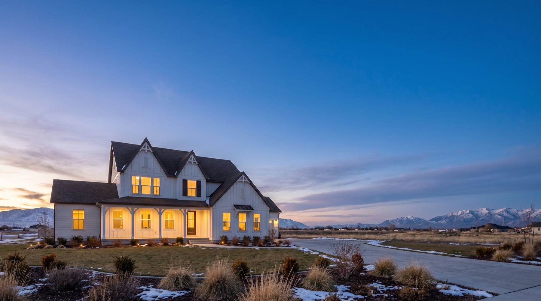 A Destination Homes Canterbury at dusk on the left of a wide evening sky, warm light in every window and the snow-dusted Wasatch behind it